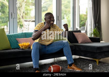 Ein glücklicher afroamerikanischer Mann, der sich das american Football-Spiel ansieht und zu Hause unterstützt Stockfoto