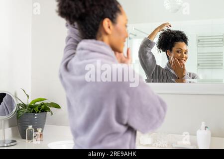 Eine glückliche, birassische Frau im Bademantel, die in den Spiegel im Badezimmer schaut Stockfoto