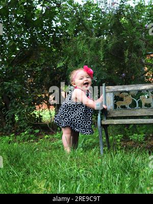 Das kleine Mädchen hält sich auf einer Holzbank fest und drückt seine Freude durch Gurgeln und Kochen aus. Sie ist barfuß und trägt ein Sonnenkleid. Rosa Blumenband umkreist Stockfoto