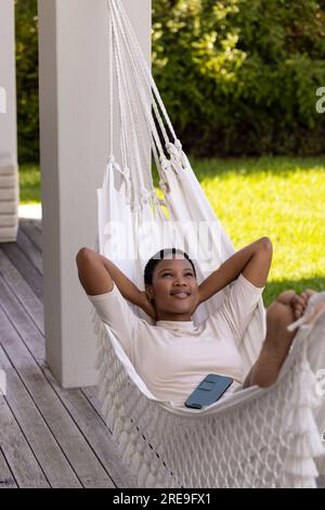 Glückliche afroamerikanische Frau mit Smartphone, die in der Hängematte auf der Terrasse lag Stockfoto