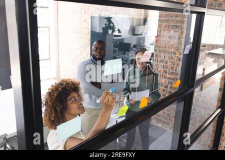 Fokussierte, vielfältige Kollegen diskutieren die Arbeit und machen sich Notizen an der Glaswand im Büro Stockfoto