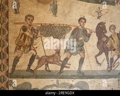 Das Mosaik im Raum der kleinen Jagd, Villa Romana del Casale, Sizilien, zeigt zwei Männer mit einem Jagdhund, der ein Eber mit einem Faden auf einer Stange trägt. Stockfoto