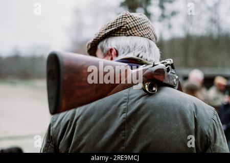 Ein älterer Mann auf der Jagd. Seniorjäger mit grauen Haaren, Tweed-Mütze und Öljacke mit einer Schusswaffe auf der Schulter. Fasanenjagd im Herbst. Stockfoto