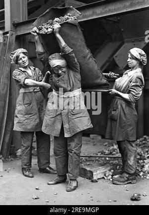 Horace Nicholls - zwei weibliche Beutelfüller verwenden zwei kurze Stangen, um einer weiblichen Koksheiterin zu helfen, während sie einen großen Sack Koks auf ihren Rücken hebt - Stockfoto