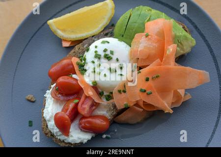 Frühstücks-, Brunch- oder Mittagssandwich mit geröstetem Roggenbrot, Frischkäse, Tomaten, gekochtem Ei, Avocado und dünn geschnittenen Karotten auf einem blauen Teller Stockfoto