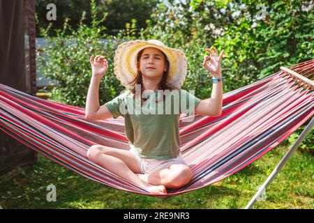 Outdoor-Aktivitäten im Sommer. Außerhalb der Stadt, Hütte. Süßes Teenager-Mädchen, das auf einer Hängematte in Lotusposition sitzt, Yoga, entspannen Stockfoto