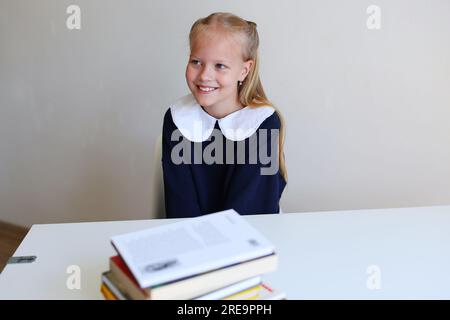 Foto eines süßen, charmanten Schulmädchens, das lächelt und an einem weißen Tisch sitzt Stockfoto