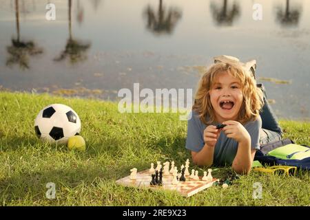 Aufgeregtes Kind, das Schachspiel im Garten spielt, auf Gras liegt. Konzentriertes Kinderspiel Schach. Kind spielt Brettspiel draußen. Stockfoto