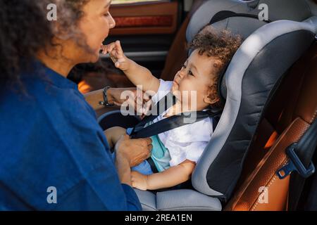 Ein kleiner Junge, der mit seiner Mutter spielt, während sie die Gurte seines Autositzes justiert Stockfoto