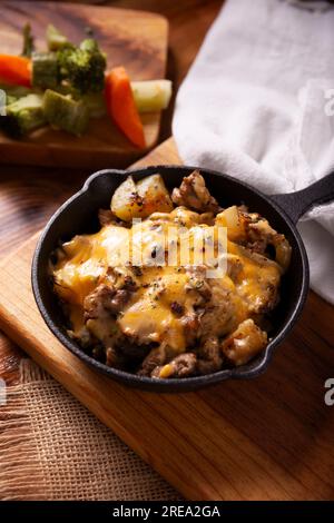 Hausgemachtes Rezept für Kartoffeln mit Steak und Schmelzkäse, gewürzt und serviert in einer gusseisernen Pfanne auf einem rustikalen Holztisch. Stockfoto