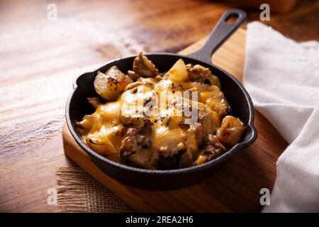 Hausgemachtes Rezept für Kartoffeln mit Steak und Schmelzkäse, gewürzt und serviert in einer gusseisernen Pfanne auf einem rustikalen Holztisch. Stockfoto