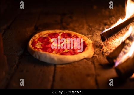 Italienische Pizza Peperoni wird in einem Holzofen zubereitet. Pizza mit Salami im heißen Ofen in der Nähe brennender Feuerwälder. Lebensmittelfotografie Stockfoto
