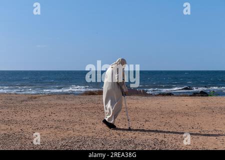 Ein Bild von einer alten Frau mit Krücke, die an der Küste entlang geht, das blaue Meer ist in einer angenehmen Atmosphäre, eine alte Frau in Bademänteln am Strand Stockfoto