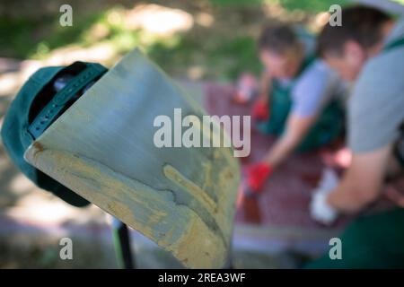 Eine Stahlschaufel, die sich vor dem Hintergrund von Arbeitern, die an den Pflastersteinen arbeiten, gegen eine andere neigt. Stockfoto