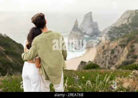 Ein Paar, das ein romantisches Date hat, ein Mann, der eine Frau umarmt, sich vor die Kamera stellt und einen wunderschönen Blick auf das Meer genießt Stockfoto