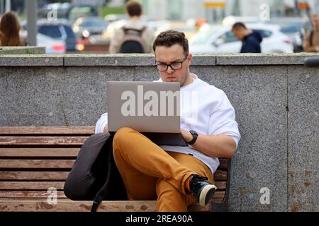 Ein Typ mit Brille, der mit einem Laptop auf einer Bank auf der Sommerstraße sitzt Stockfoto