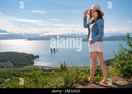 Hipster-Frau, die in die Kamera schaut, mit Strohhut, Jeansjacke, weißen Shorts und Sandalen. Eine Frau, die auf einem Berg im Hintergrund des Ozeans steht Stockfoto