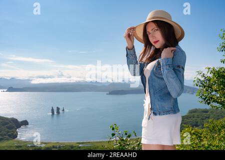 Erwachsene Hipster-Frau mit Strohhut, Jeansjacke und weißen Shorts, die am Hochsitz stehen und am sonnigen Sommertag einen Panoramablick auf das Meer genießen Stockfoto