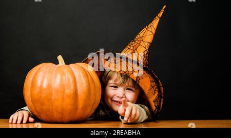 Ein lächelnder Junge mit Hexenhut und halloween-Kürbis, der auf dich zeigt. Süßes oder Saures. Kleiner Junge mit Kürbis. Vorbereitung auf den Halloween-Urlaub. Glücklich Stockfoto