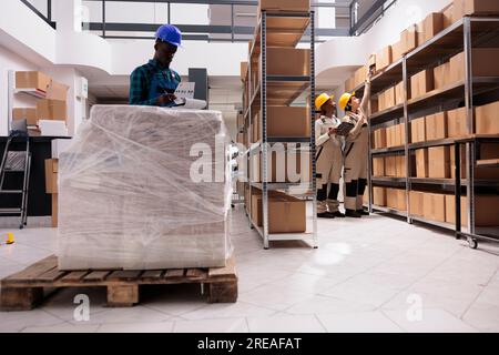 Ein Lagerverwalter aus Afrika überprüft die Paketinformationen in der Zwischenablage, verwaltet die Frachtsendung. Verschiedene Mitarbeiter von Versandunternehmen, die im Industrielager arbeiten Stockfoto