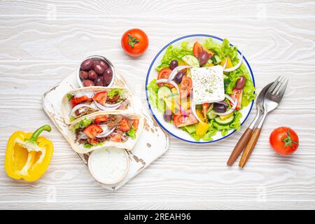 Traditionelle griechische Speisen: Griechischer Salat, Gyros mit Fleisch und Gemüse, Tzatziki-Sauce, Oliven auf weißem rustikalem Holztisch von oben Stockfoto