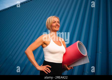 Porträt einer mittelalterlichen wunderschönen blonden Frau mit Sportmatte auf blauem Hintergrund. Gesunder, aktiver Lebensstil, Yoga-Workout Stockfoto