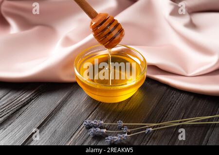 Fließender Honig. Bio-Honig wird aus einem Holzstiel in eine transparente Schüssel auf Holz- und Textilhintergrund gegossen. Stockfoto