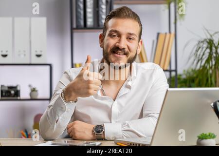 Wie. Glücklicher Geschäftsmann aus dem Nahen Osten, der an einem Laptop arbeitet, guckt zustimmend auf die Kamera und zeigt Daumen nach oben, wie ein positives Zeichen für etwas Gutes, positives Feedback am Arbeitsplatz. Freiberufler Stockfoto
