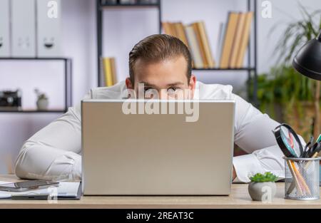 Ein selbstbewusster libanesischer Geschäftsmann, der sich hinter einem Laptop versteckt, in die Kamera schaut, seine Kollegen bei der Arbeit ausspioniert, piepst. Professioneller Freiberufler, der von hinten mit gerissenen Augen auf den Computer blickt Stockfoto