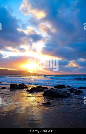 Dunkle nasse große Steine am Strand am Wasserrand bei Sonnenuntergang. Meereslandschaft mit schweren dunklen Wolken und untergehender Sonne. Stockfoto