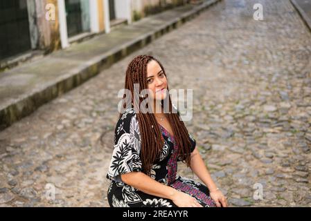 Eine Frau hockte auf einer Kopfsteinpflasterstraße. Haargeflechte und afrikanisches Kleid. Pelourinho, Brasilien. Stockfoto