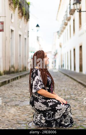 Eine Frau hockte auf einer Kopfsteinpflasterstraße. Haargeflechte und afrikanisches Kleid. Pelourinho, Brasilien. Stockfoto