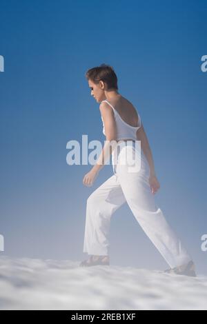 Schlanke blonde Frau läuft durch Schnee mit leichtem Nebel bei sonnigem Wetter und blauem Himmel. Hübsche Erwachsene Frau mit kurzen Haaren in weißer Hose Stockfoto