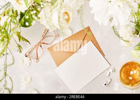 Feminine flache Lage mit handgemachtem pastellfarbenem Papierumschlag für Gruß, Einladung, Text in weißen Blumen. Gläser, Teetasse und Ohrhörer, Draufsicht. Stockfoto