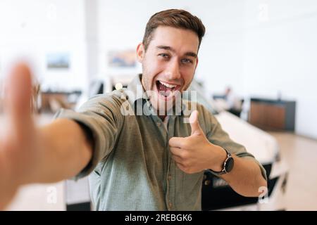 POV-Bild eines fröhlichen Käufers, der nach dem Kauf eines neuen Fahrzeugs ein Selfie mit Daumenschild auf dem Smartphone macht. Ein lächelnder junger Mann Stockfoto