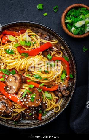 Bratnudeln mit Gemüse umrühren: Rote Paprika, Champignons, grüne Zwiebel und Sesamsamen in Keramikschüssel. Schwarzer Tischhintergrund, Draufsicht Stockfoto