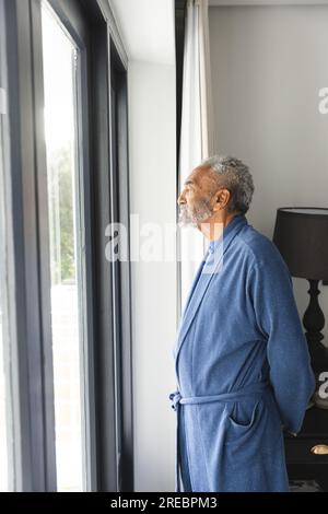 Rücksichtsvoller, birassistischer Senior-Mann, der Bademantel trägt und zu Hause durch das Fenster schaut Stockfoto