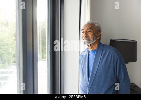 Rücksichtsvoller, birassistischer Senior-Mann, der Bademantel trägt und zu Hause durch das Fenster schaut Stockfoto