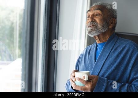 Rücksichtsvoller älterer, birassistischer Mann, der Bademantel trägt und zu Hause eine Tasse Kaffee am Fenster hält Stockfoto