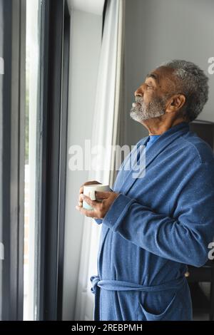 Rücksichtsvoller älterer, birassistischer Mann, der Bademantel trägt und zu Hause eine Tasse Kaffee am Fenster hält Stockfoto