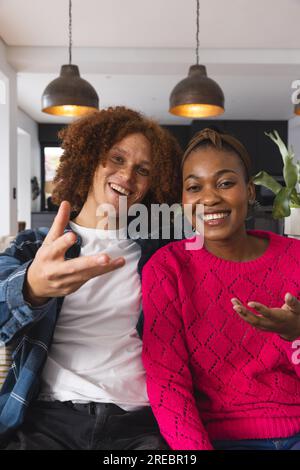 Glückliches, vielseitiges Paar, das Videoanrufe führt, zusammen auf dem Sofa sitzt und zu Hause Gesten macht Stockfoto