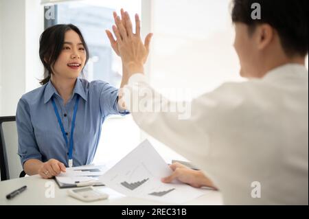 Eine fröhliche junge asiatische Geschäftsfrau oder Büroangestellte gibt einem männlichen Kollegen ein Happy Five, während sie gemeinsam an einem Projekt arbeiten. Teamwork, Co-wo Stockfoto
