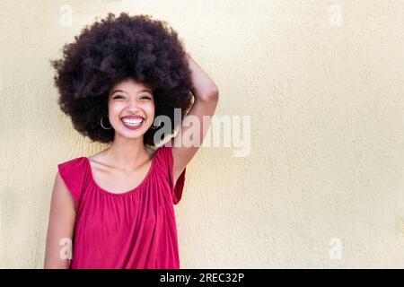 Porträt einer positiven afroamerikanischen Frau mit Afro-Haarschnitt, die mit erhobener Hand lächelt und in die Kamera schaut, während sie gegen beigefarbene Hintergrunen steht Stockfoto