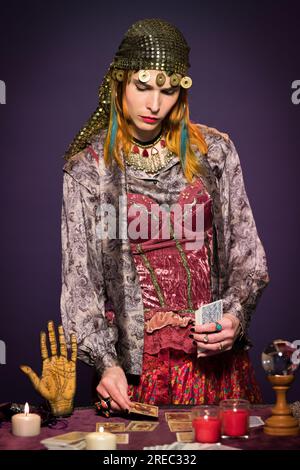 Eine seriöse junge Wahrsagerin mit langen Ingwerhaaren in Bo-Style-Kleidung, die Tarotkarten auslegt, die am Tisch stehen, mit brennenden Kerzen und Schreien Stockfoto