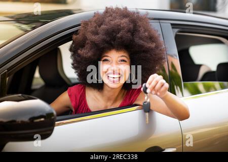 Glückliche afroamerikanische Frau in Afro-Haar und Make-up lächelt und schaut in die Kamera, während sie im neuen Auto sitzt und den Autoschlüssel vorführt Stockfoto