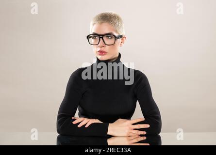 Seriöses weibliches Modell mit schwarzem Rollkragenpullover und Brille kreuzt die Arme, während es auf einem Spiegeltisch auf grauem Hintergrund sitzt und wegschaut Stockfoto
