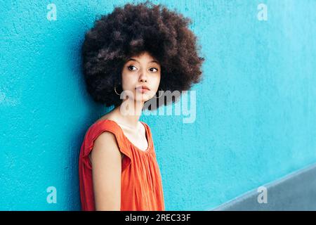 Seitliches Porträt einer jungen marokkanischen Frau in legerer Kleidung mit Afro-Haarschnitt, die auf die Kamera schaut und vor blauem Hintergrund steht Stockfoto