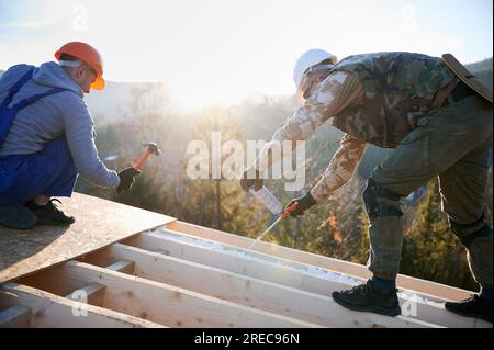 Männlicher Baumeister, der am Morgen Wärmedämmung auf dem Dach des Holzrahmenhauses durchführt. Männlicher Arbeiter, der Polyurethanschaum auf das Dach des zukünftigen Cottage spritzt. Bau- und Dämmkonzept. Stockfoto