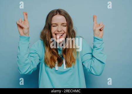 Fröhliches, verrücktes Teenager-Mädchen mit gewelltem Omelhaar, Augen geschlossen, Daumen gekreuzt, Zunge herausragt, steht an der blauen Wand im Studio, sorgenfrei und h Stockfoto