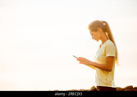 Nahaufnahme einer jungen Frau in Sportbekleidung, die ihr Smartphone in der Hand hält, nimmt zwischen den Workouts Selfie vor dem Hintergrund eines orangefarbenen Morgenhimmels auf. Das c Stockfoto